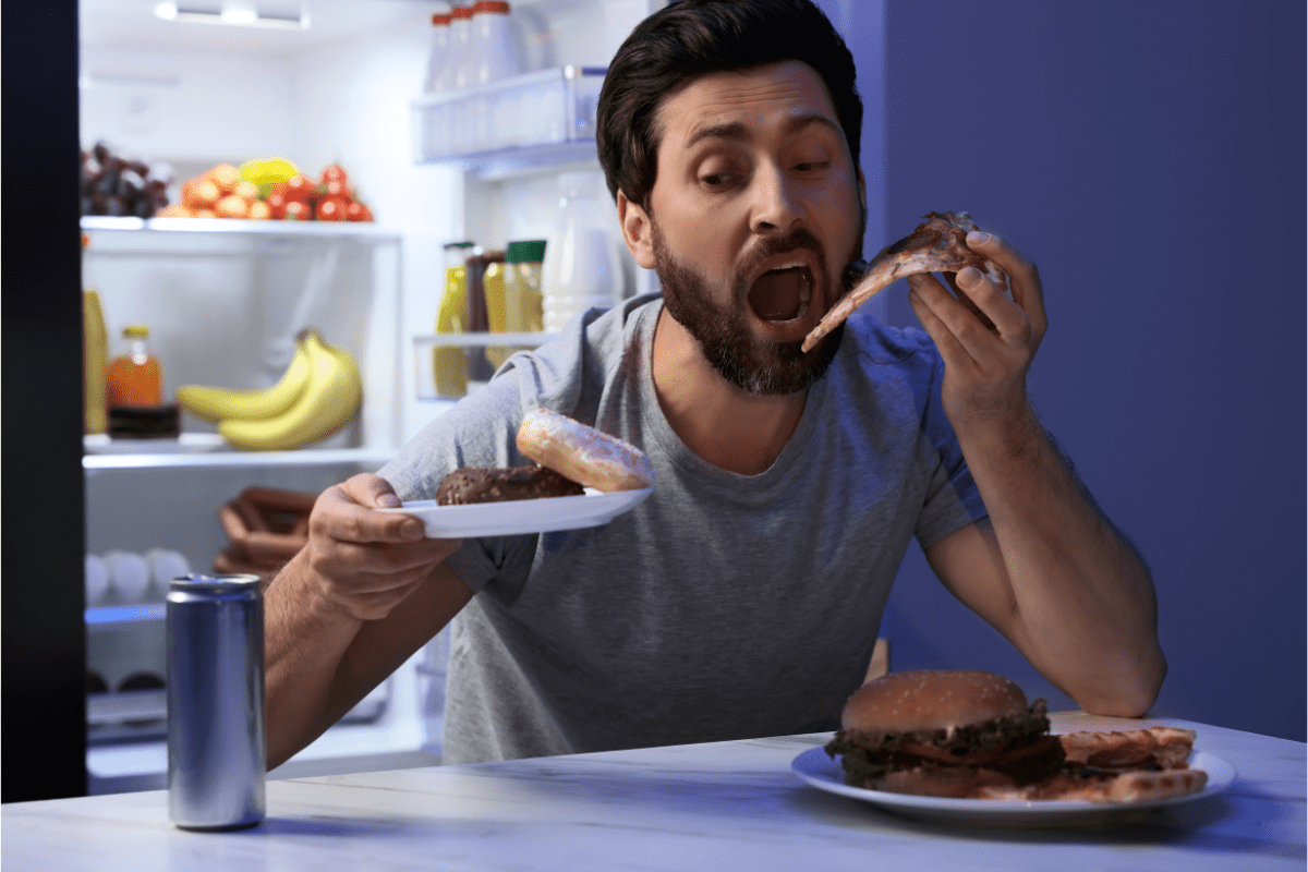 Man eating pizza, burgers, and sweets late at night in front of an open fridge.