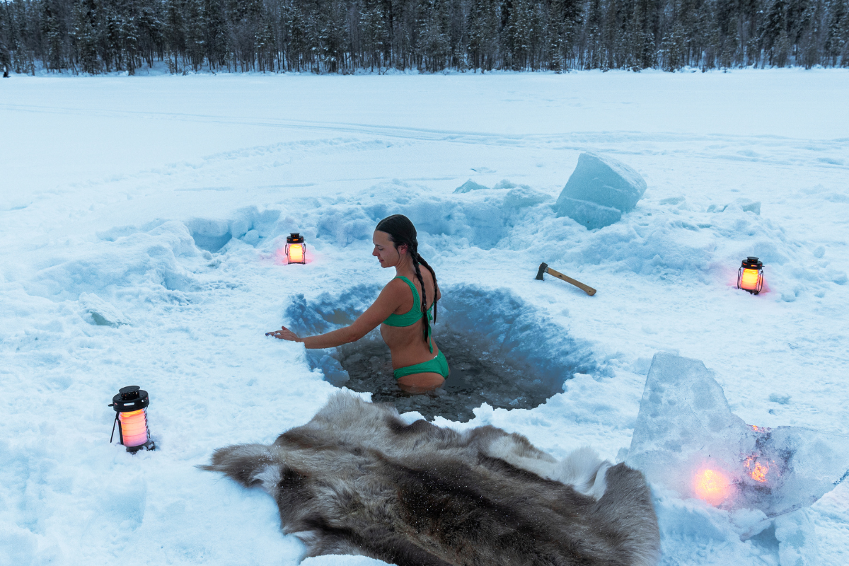 A person in a green bikini is seen immersing themselves in an ice hole surrounded by snow.