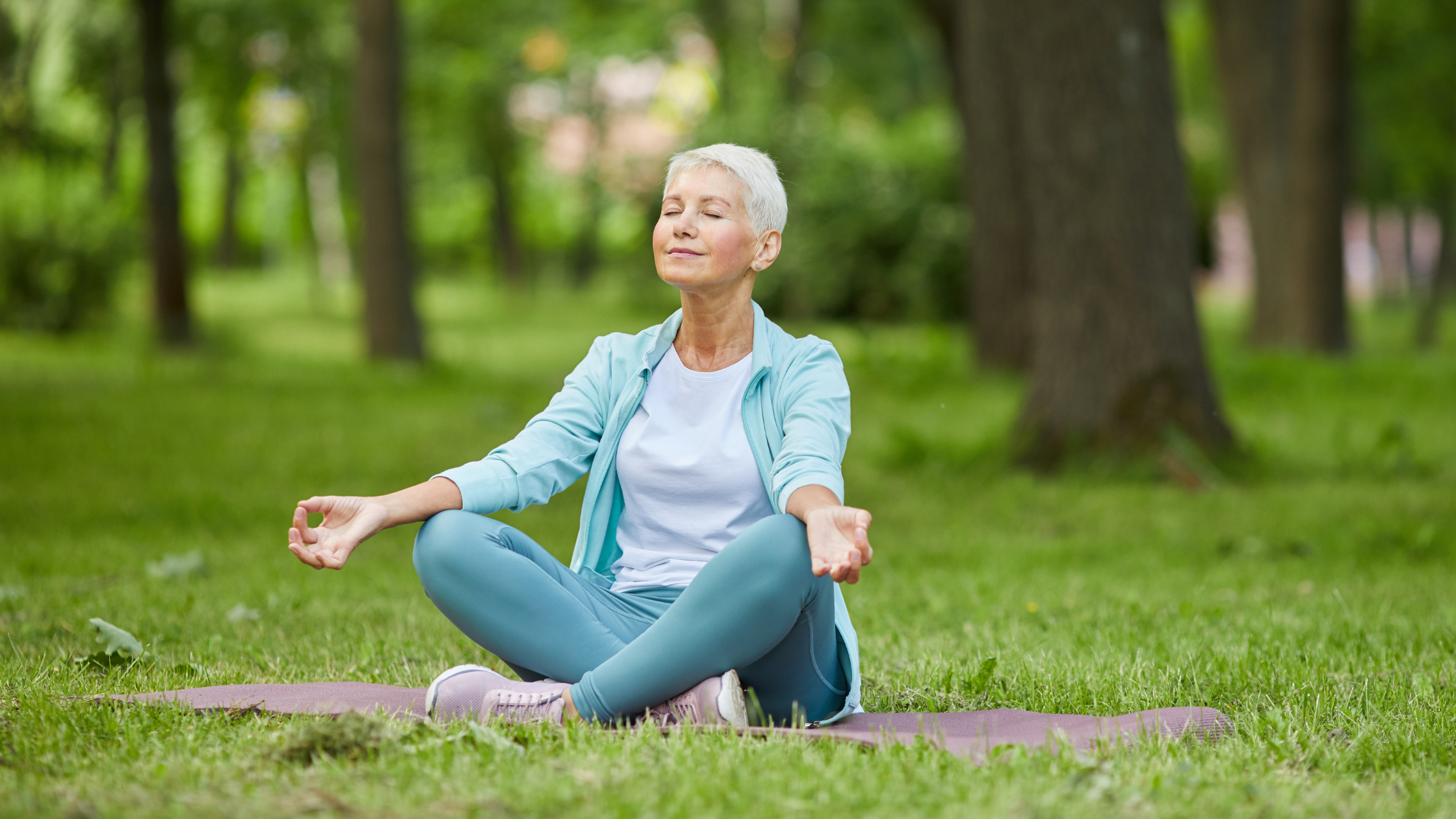 A woman practising meditation and stretching in a serene outdoor setting, sitting cross-legged on a yoga mat with her eyes closed, exuding a sense of calm and relaxation.