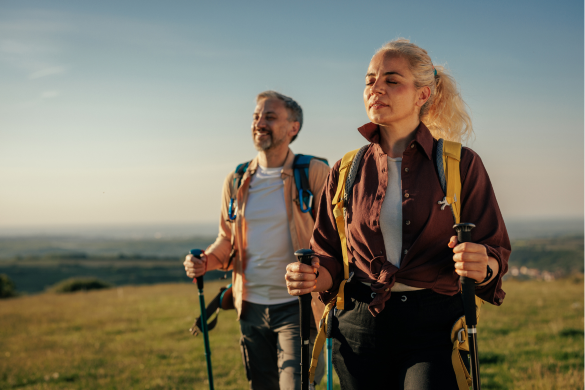Two older adults, a man and a woman, enjoy a hike in a scenic landscape, equipped with hiking poles and backpacks.