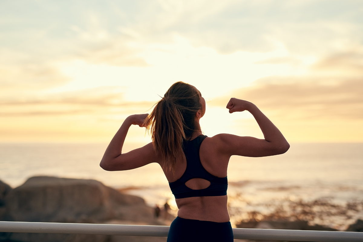Woman flexing arms at sunrise by the ocean.