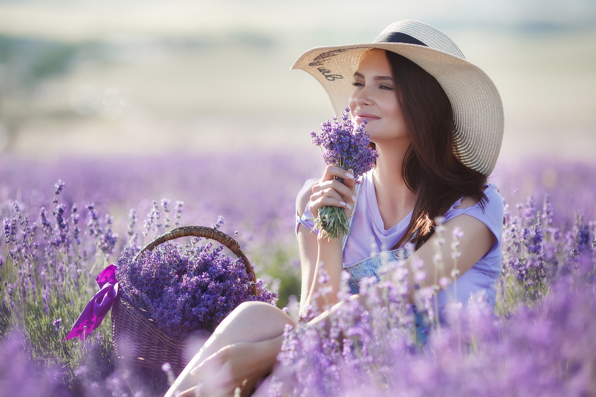 A woman in a straw hat sits in a lavender field, holding a bouquet and smiling. Beside her is a basket filled with lavender flowers, tied with a purple ribbon.