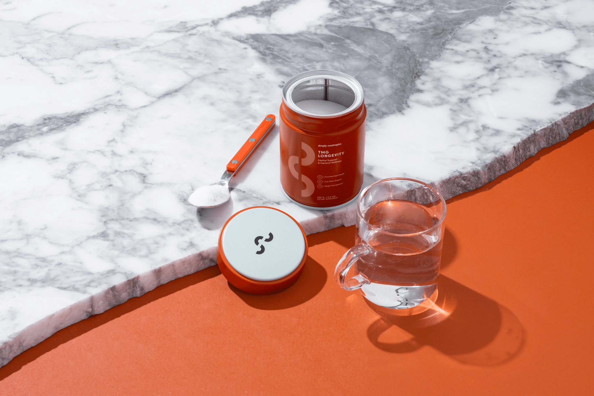 Simply Nootropics TMG Powder on a marble surface with a scoop of powder, lid, and a glass of water nearby.