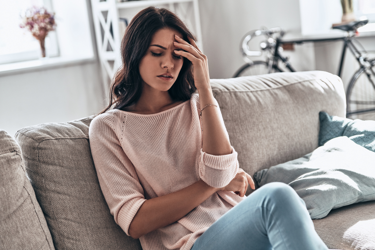 Woman sitting on a sofa with her hand on her forehead, looking tired or unwell.