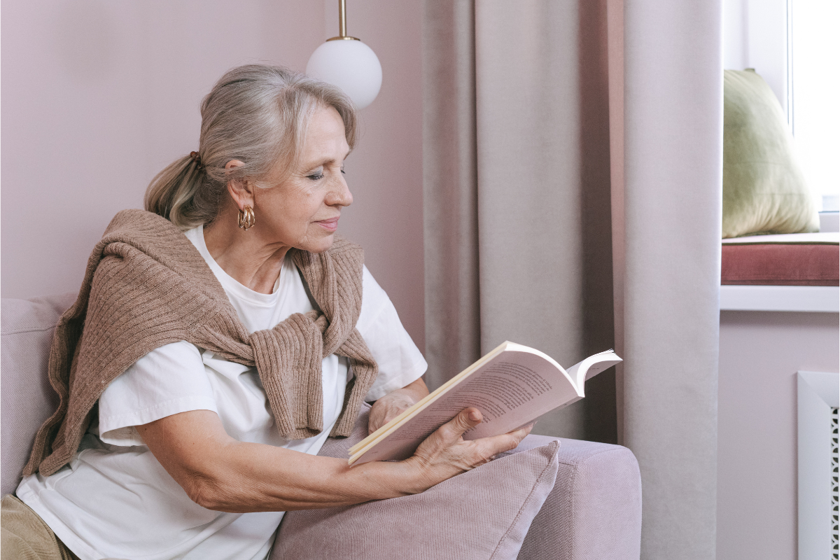Elderly woman reading a book on a cosy pink couch.