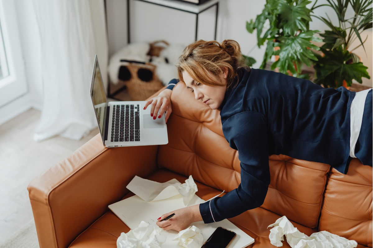 Woman resting head on laptop while sitting on couch with tissues nearby.