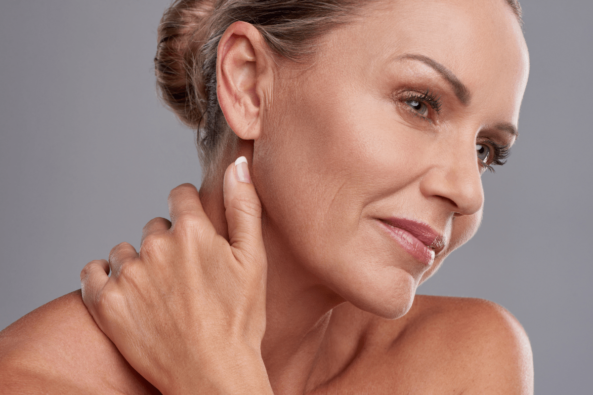 Close-up of a smiling woman with radiant skin, gently touching her neck and shoulder.