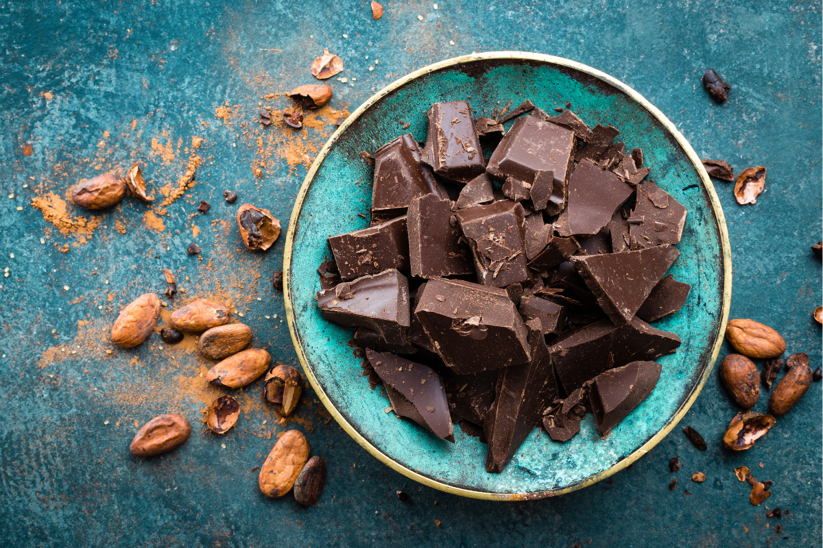 A vibrant teal bowl filled with chunks of rich, dark chocolate sits on a textured blue surface, surrounded by scattered cocoa beans and cocoa powder.