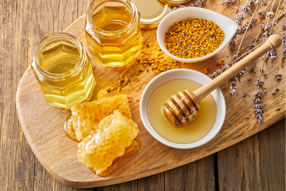 Assorted jars and bowls of honey, honeycomb, and bee pollen displayed on a wooden board.