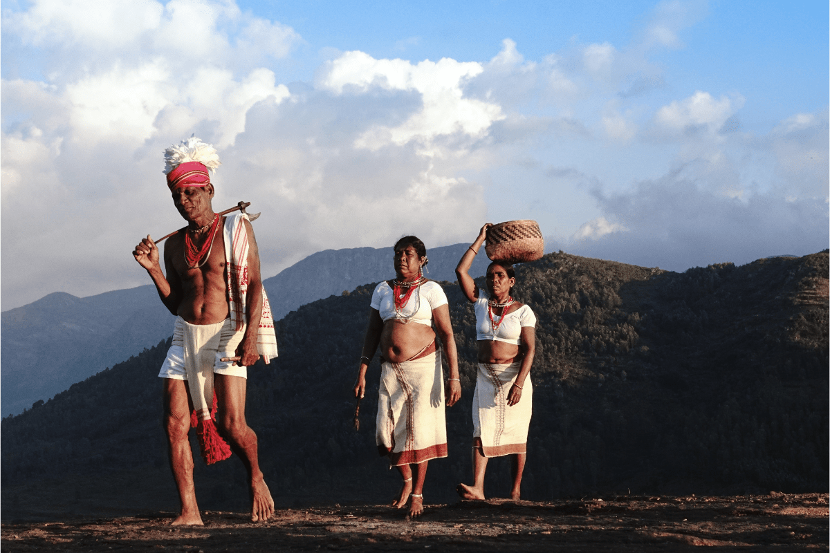 Three Indigenous people in traditional attire walk barefoot through a hilly landscape.