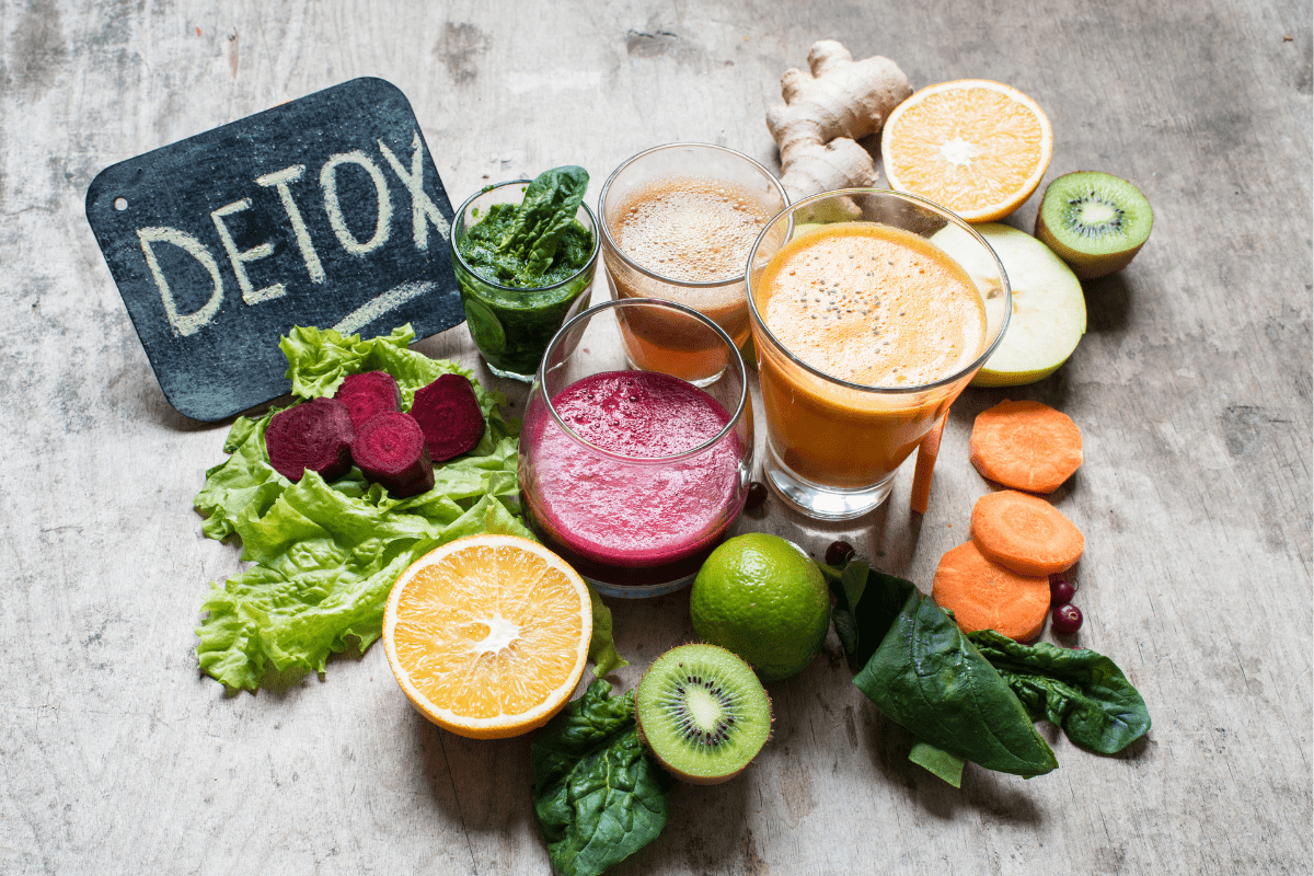 Fresh detox ingredients including juices, leafy greens, citrus fruits, and root vegetables on a wooden table with a 'Detox' sign.