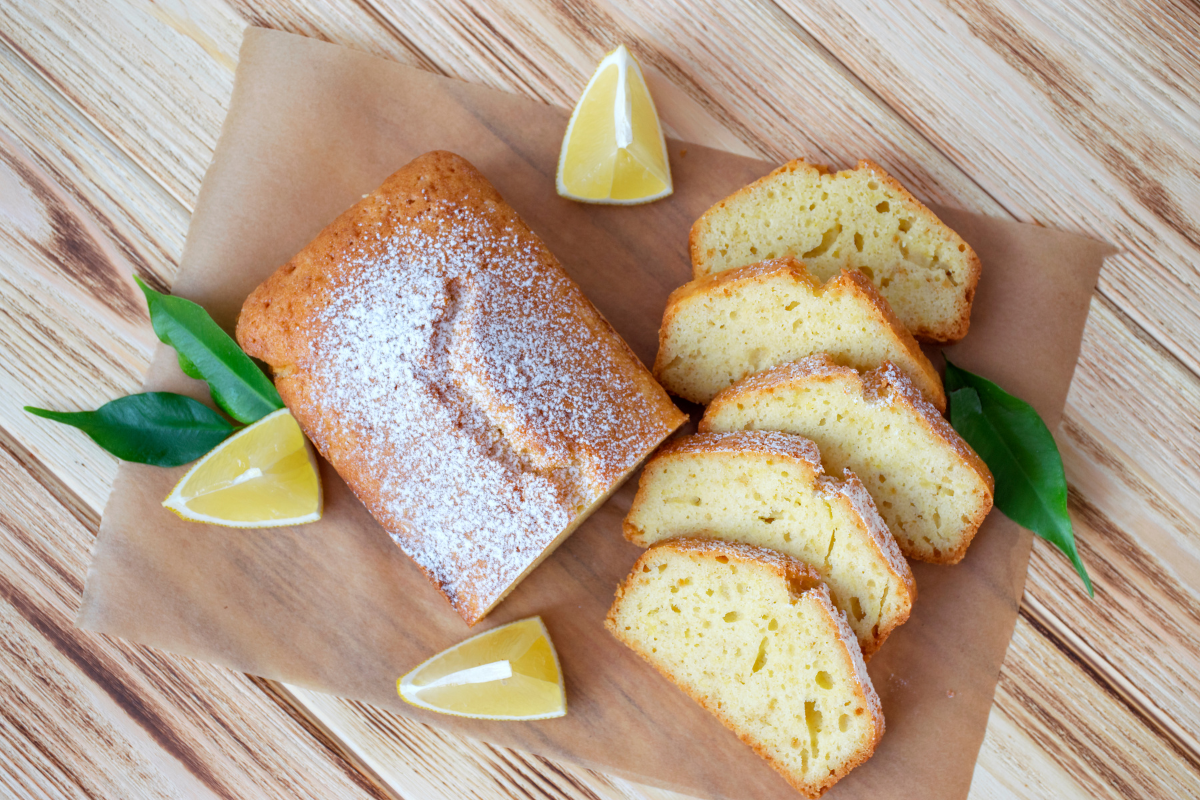 Lemon loaf cake dusted with icing sugar on brown paper with lemon wedges.