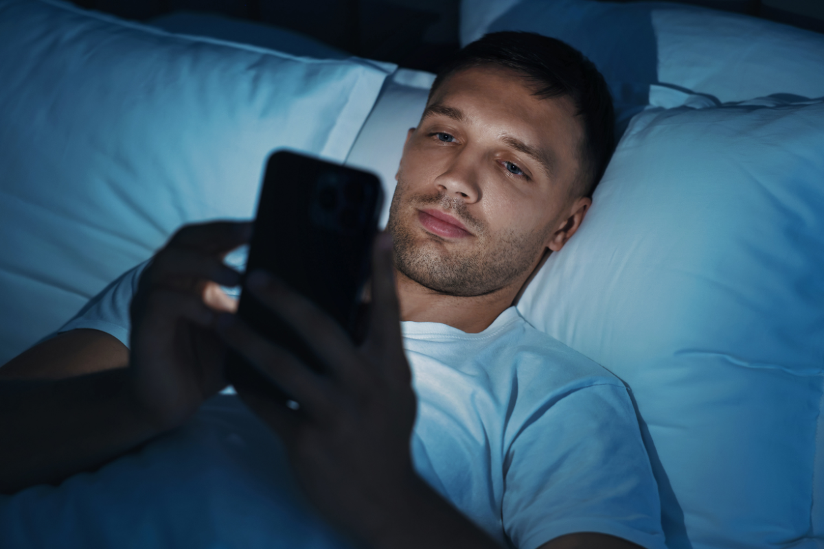 Man lying in bed at night using his phone, lit by blue screen light.