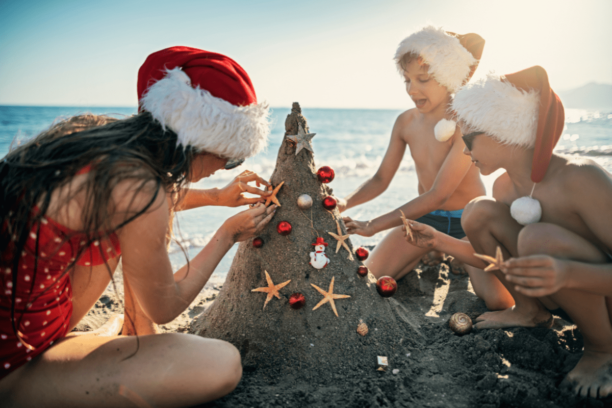 Children and a woman decorating a sandcastle with holiday ornaments on a sunny beach, wearing Santa hats.