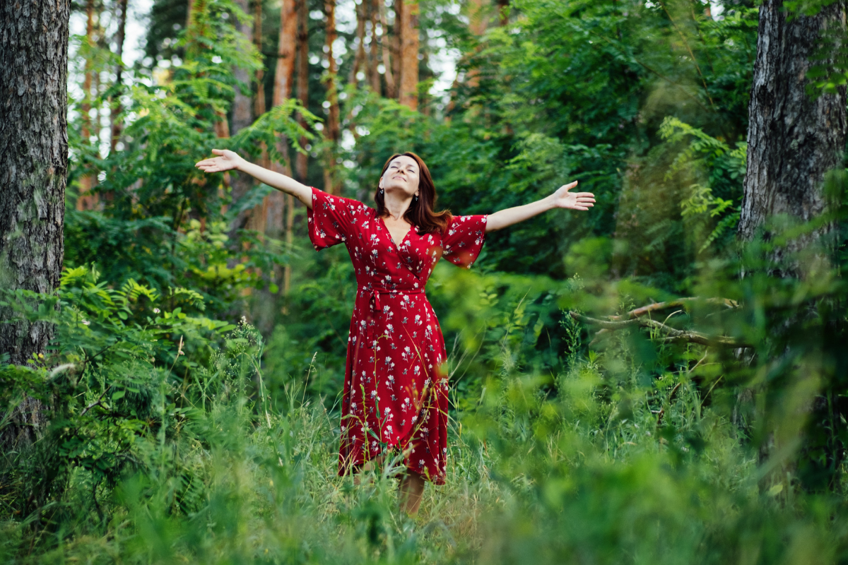 Woman in a red floral dress standing in a lush green forest, arms outstretched, enjoying nature.