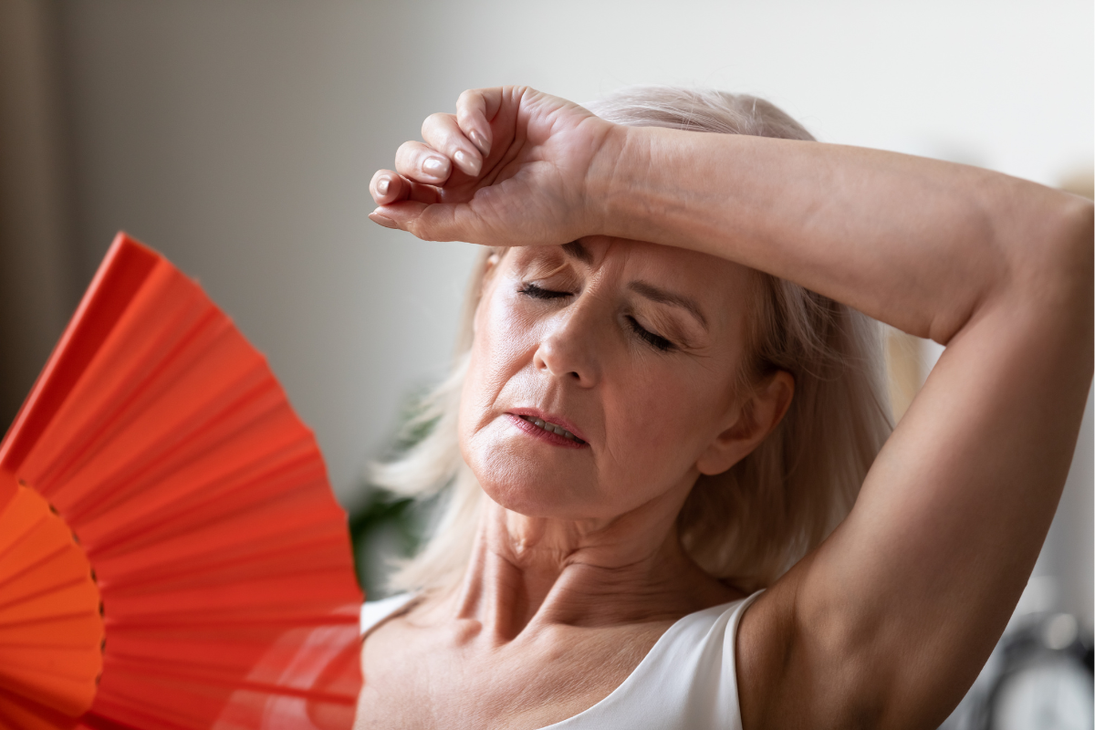 A middle-aged woman with her eyes closed, resting her forehead on her forearm, holds a red fan, suggesting she is experiencing a hot flash or discomfort.