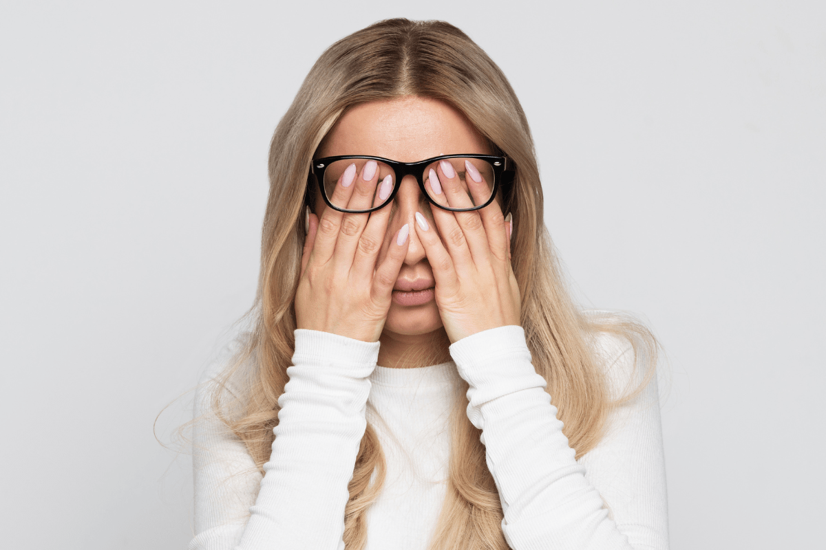 A woman in glasses covering her face with her hands, expressing fatigue or stress.