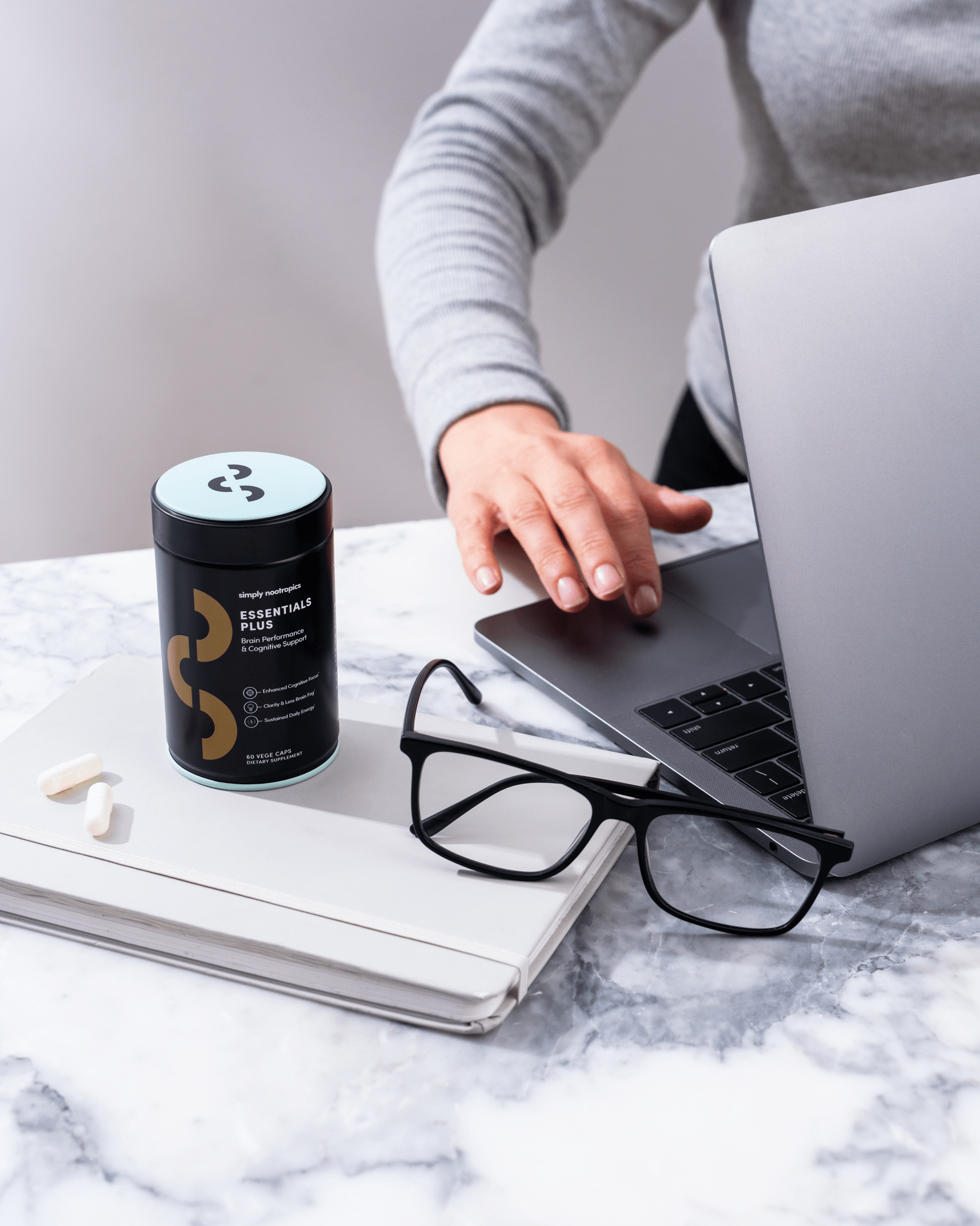 Person reaching for laptop beside a container of Simply Nootropics Essentials Plus, glasses, and notebook on a marble surface.