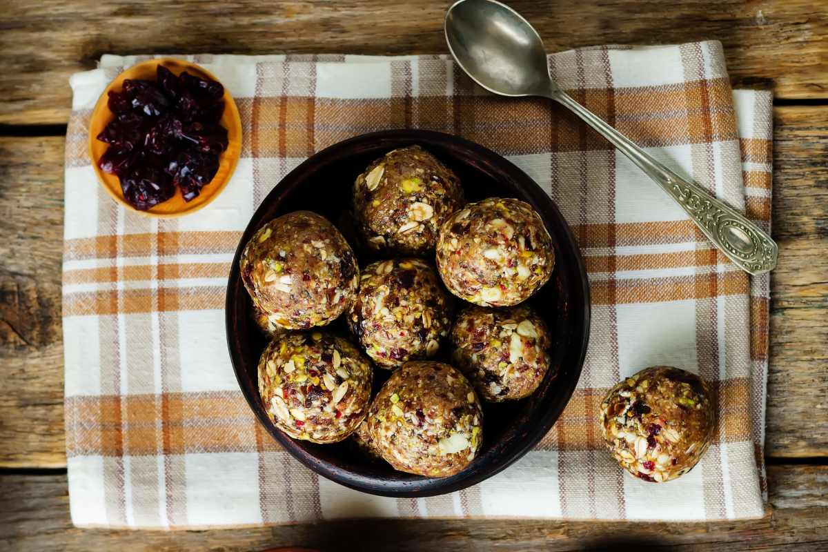 Bowl of oat and nut energy bites on a checkered cloth with dried cranberries and a spoon.