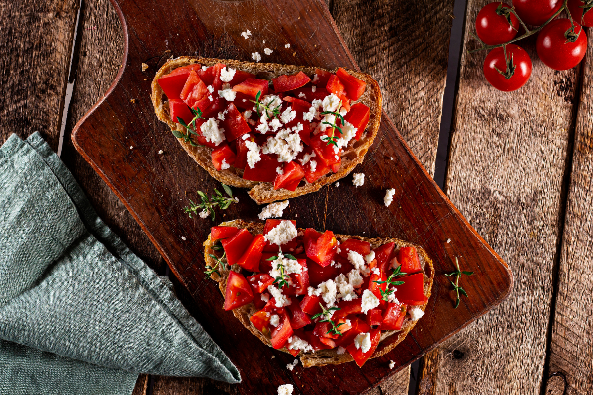 Two slices of toasted bread topped with chopped tomatoes, feta cheese, and fresh herbs on a wooden board.