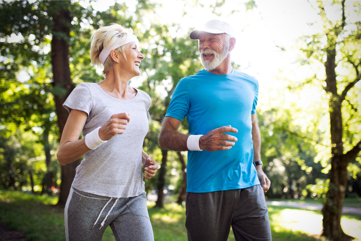 A senior couple jogging and smiling in a sunlit park, enjoying outdoor exercise.