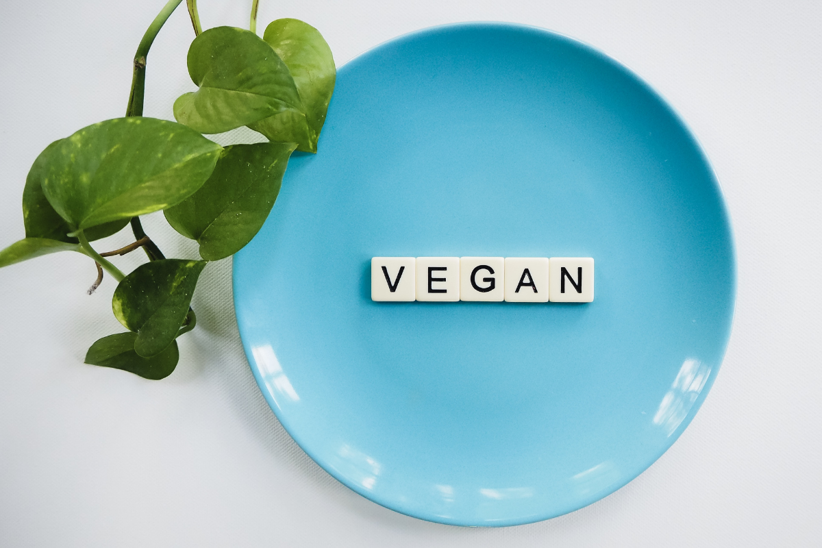 Blue plate with the word “VEGAN” spelled in tiles, beside green leaves.