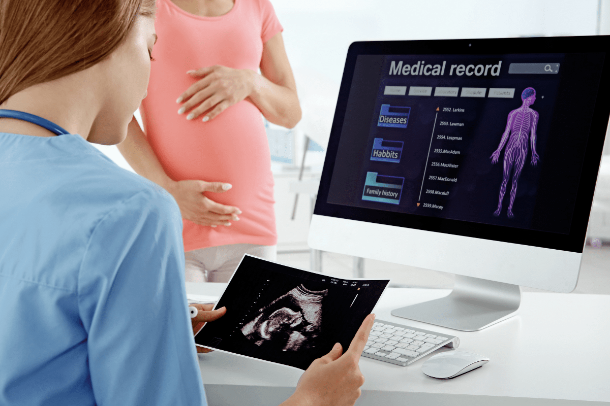 A medical professional reviews an ultrasound image on a tablet while a pregnant patient stands nearby, holding her stomach.