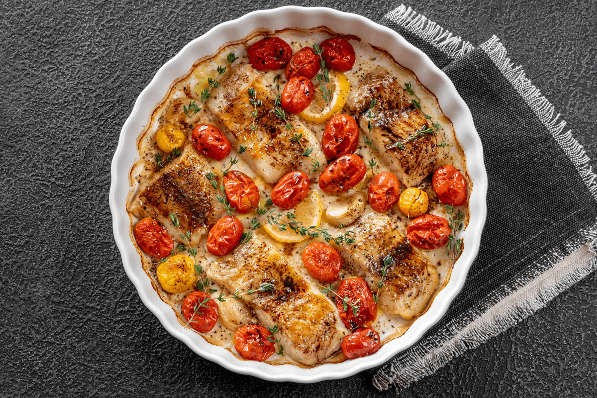 Baked cod with cherry tomatoes and lemon slices in a white dish, garnished with herbs.