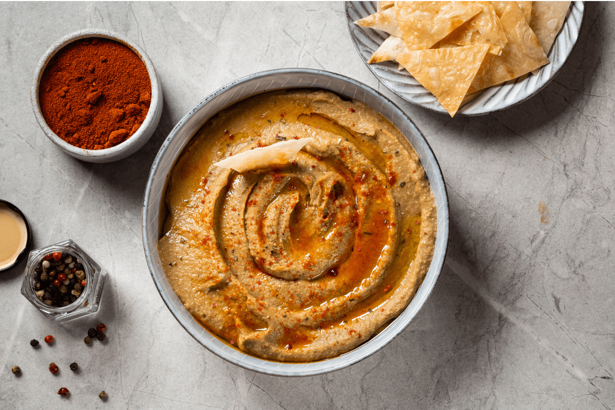 A bowl of creamy roasted eggplant and garlic dip, drizzled with olive oil and sprinkled with spices, served alongside crispy pita chips.