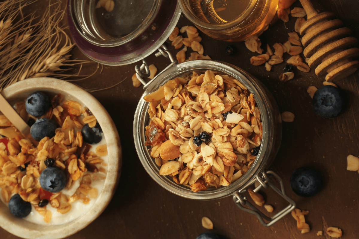 A jar of homemade granola, surrounded by fresh blueberries, a bowl of yoghurt topped with granola and berries, and a drizzle of honey.