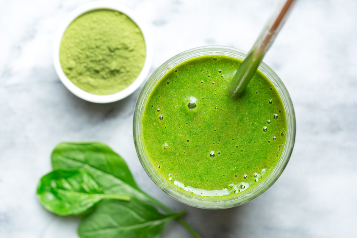 Top view of a green smoothie in a glass with a metal straw, surrounded by fresh spinach leaves and a bowl of green powder.