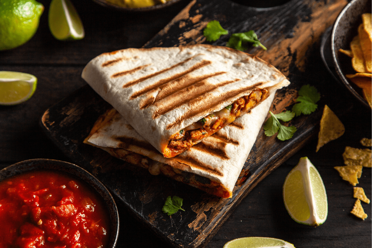 Grilled quesadilla filled with a savoury mixture, served with lime wedges and salsa on a rustic board.