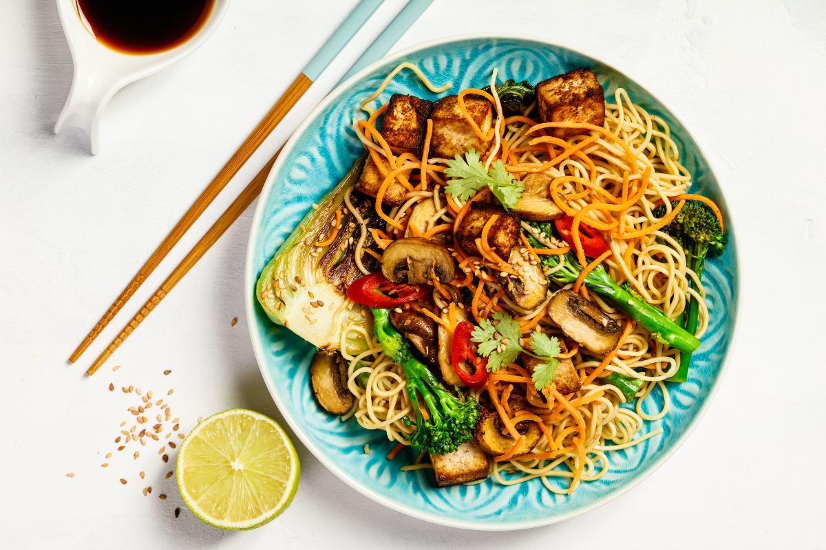 A vibrant bowl of stir-fry noodles with broccoli, mushrooms, bell peppers, and tofu, garnished with sesame seeds and fresh cilantro, served with a slice of lime.