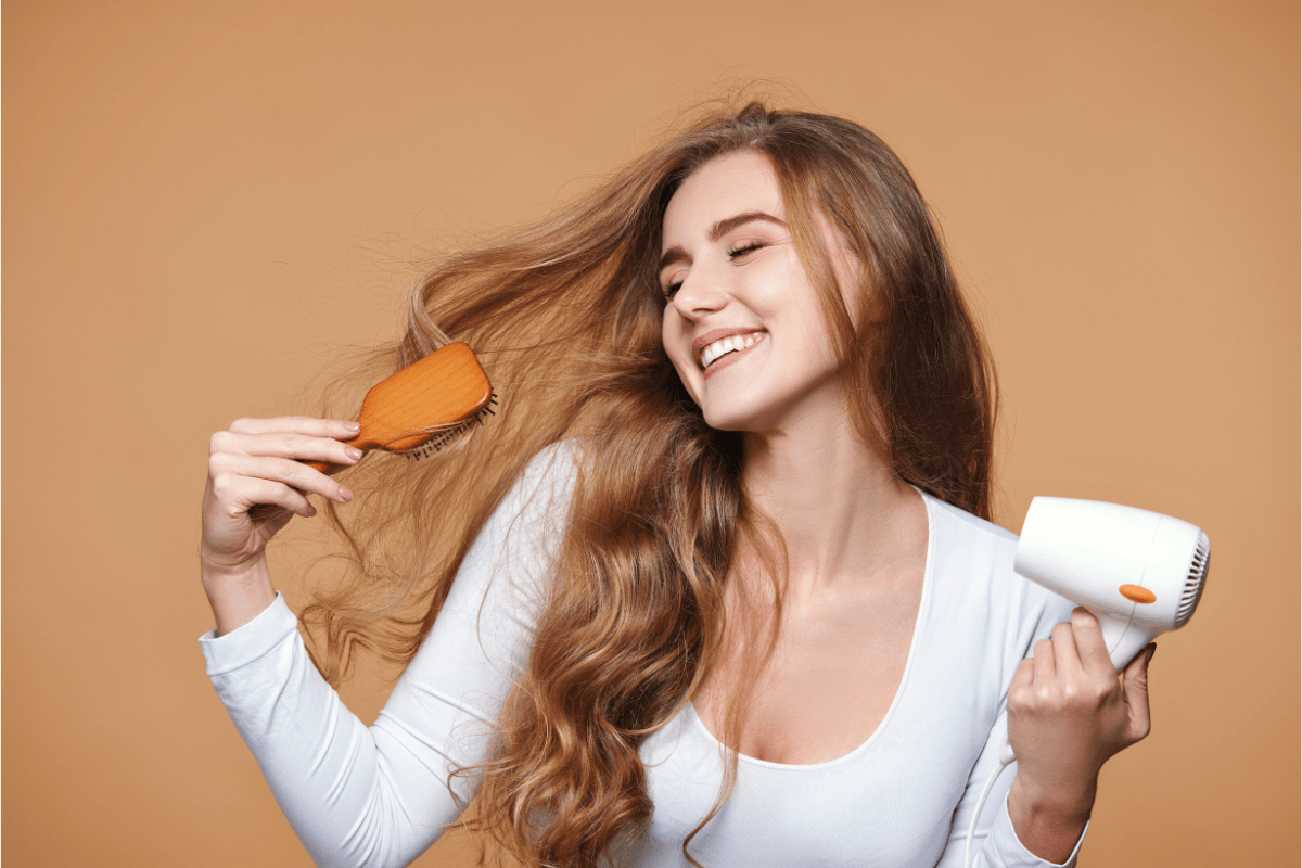 Smiling woman brushing her long hair while holding a hairdryer, on a beige background.