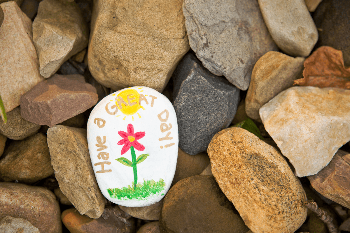 Painted rock with a flower and the words 'Have a Great Day!' among natural stones.